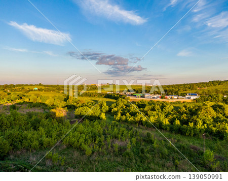Aerial view of gas fuel station near road. Green trees and bushes in foreground during the sunset. Blue sky with clouds Aerial view of gas fuel station near road. Green trees and bushes in foreground during the sunset. Blue sky with clouds 139050991