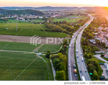 Aerial above view of highway road with cars. Highway leads from bottom to top through the trees and bushes. City panorama with buildings in background. Sunny day, sunset Aerial above view of highway road with cars. Highway leads from bottom to top through the trees and bushes. City panorama with buildings in background. Sunny day, sunset 139050996