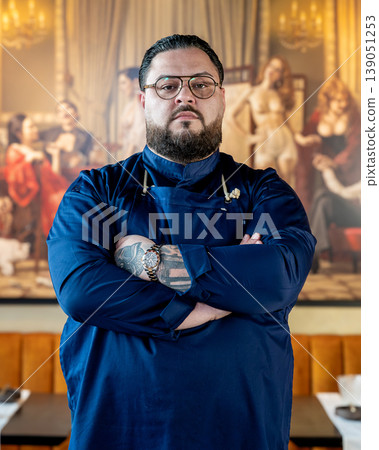 Chef portrait in restaurant, close cropped detail showing folded arms and tattoos, navy jacket texture and calm intensity highlight Chef portrait in restaurant, close cropped detail showing folded arms and tattoos, navy jacket texture and calm intensity highlight 139051253