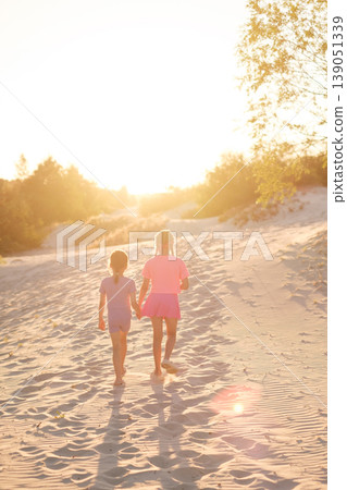 The girls of the sisters walk along the sandy road towards sunset. Vertical photo. Harmony and tranquility. The girls of the sisters walk along the sandy road towards sunset. Vertical photo. Harmony and tranquility. 139051339