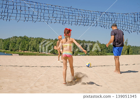 A father and his daughters are playing beach volleyball on the shore of a lake. A homemade net for playing volleyball. Kids in swimsuits having fun with dad A father and his daughters are playing beach volleyball on the shore of a lake. A homemade net for playing volleyball. Kids in swimsuits having fun with dad 139051345