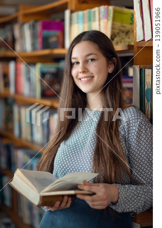 Young smiling girl reading a look. Student with an open book standing near shelves in library, choosing literature for reading 139051966