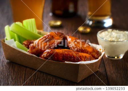 Buffalo BBQ chicken wings with blue cheese sauce and beer with of celery sticks in a paper container on a wooden table.Buffalo chicken wings in a disposable tray and beer on a wooden table Buffalo BBQ chicken wings with blue cheese sauce and beer with of celery sticks in a paper container on a wooden table.Buffalo chicken wings in a disposable tray and beer on a wooden table 139052233