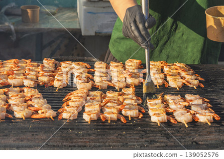 Shrimp skewers grilling on barbecue grill, chef brushes sauce, close view of seafood cooking process with rows of shrimp turning golden on hot surface. Shrimp grilling, street food barbecue 139052576