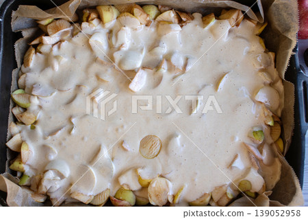 Apple pie batter poured over sliced apples in a baking pan, with rustic background, alongside fresh apples and walnuts. 139052958