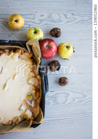 Apple pie batter poured over sliced apples in a baking pan, with rustic background, alongside fresh apples and walnuts. Apple pie batter poured over sliced apples in a baking pan, with rustic background, alongside fresh apples and walnuts. 139052960