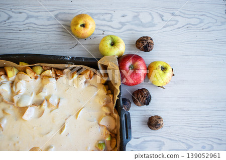 Apple pie batter poured over sliced apples in a baking pan, with rustic background, alongside fresh apples and walnuts. Apple pie batter poured over sliced apples in a baking pan, with rustic background, alongside fresh apples and walnuts. 139052961
