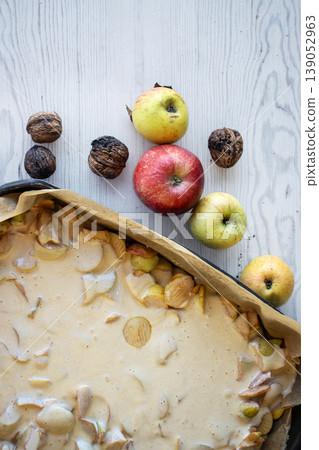 Apple pie batter poured over sliced apples in a baking pan, with rustic background, alongside fresh apples and walnuts. Apple pie batter poured over sliced apples in a baking pan, with rustic background, alongside fresh apples and walnuts. 139052963