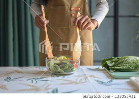A woman in an apron preparing a fresh salad on the table in kitchen. 139053431