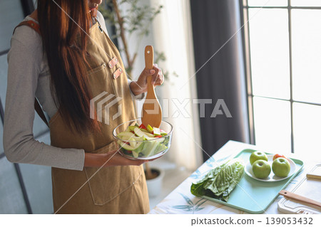 A woman in an apron preparing a fresh salad on the table in kitchen. 139053432