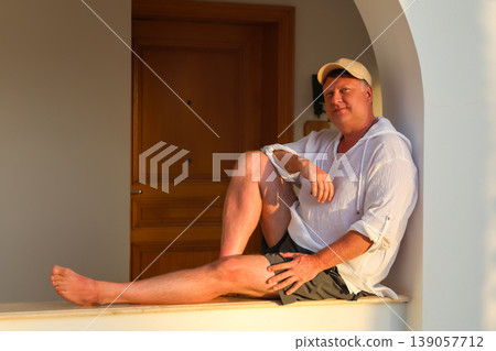 Adult man in a white shirt and cap sitting on a wide windowsill, enjoying the beautiful sunset, reflecting on life and tranquility in the golden hour 139057712