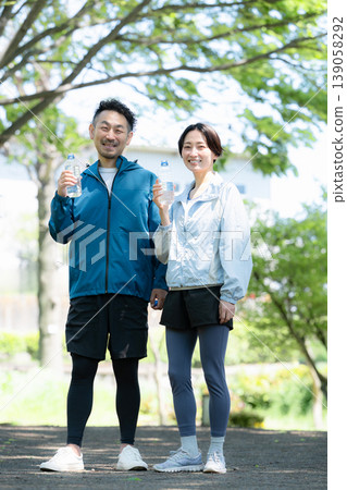 A couple, drank water from a plastic bottle while taking a break from sports outdoors amidst the fresh greenery, posing for the camera with smiles and looking directly at the camera. 139058292