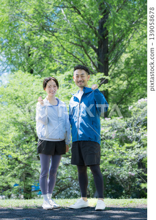 A couple, smiling and looking directly at the camera, are seen cheering on their sporting partner, who is giving a thumbs-up against a backdrop of fresh greenery. 139058678