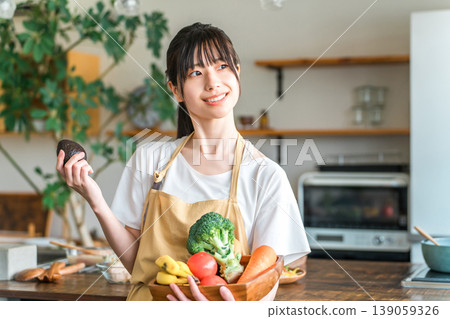 穿著圍裙的年輕亞洲女性在廚房裡拿著蔬菜和酪梨 139059326