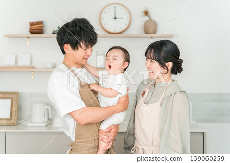 一對穿著圍裙的夫婦抱著嬰兒在廚房裡,一家人 一對穿著圍裙的夫婦抱著嬰兒在廚房裡,一家人 139060259