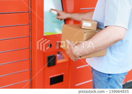 A man with cardboard boxes uses the touchscreen of an automated orange mailbox. Concept for delivery, self-service, and order pickup in e-commerce. A man with cardboard boxes uses the touchscreen of an automated orange mailbox. Concept for delivery, self-service, and order pickup in e-commerce. 139064835