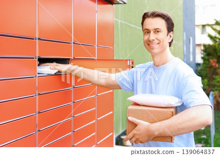 Happy young man smiling at the camera while picking up or sending packages at an automated orange parcel locker outdoors. Convenient e-commerce and self-service delivery concept. Happy young man smiling at the camera while picking up or sending packages at an automated orange parcel locker outdoors. Convenient e-commerce and self-service delivery concept. 139064837