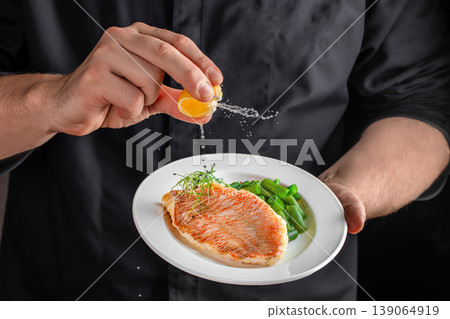 Chef squeezing lemon over grilled fish fillet with vegetables in restaurant kitchen close up 139064919