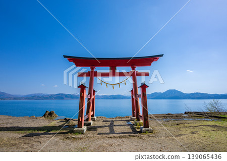 The torii gate of Gozaishi Shrine, standing on the shores of Lake Tazawa in Akita, and the breathtaking view of Lake Tazawa's blue waters reflected on the lake's surface. The torii gate of Gozaishi Shrine, standing on the shores of Lake Tazawa in Akita, and the breathtaking view of Lake Tazawa's blue waters reflected on the lake's surface. 139065436