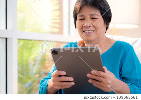 Senior asian woman relax on sofa reading tablet in living room at home. Senior asian woman relax on sofa reading tablet in living room at home. 139065661