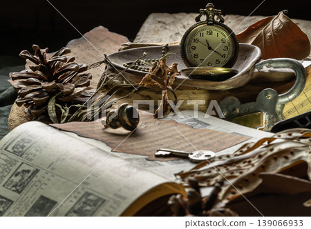 The prominent golden pocket watch serves as the focal point, resting in a wooden spoon where its brassy sheen contrasts with surrounding dried brown leaves. An open volume of aged illustrations 139066933