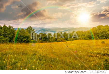 sunset over mountainous rural landscape in summertime. picturesque highland woods and grassy meadow. view of countryside scenery with forested hills behind the pasture under rainbow 139067462