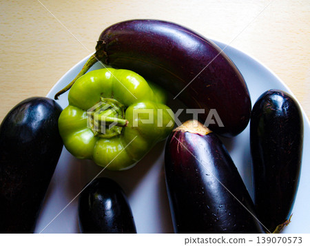 Green bell pepper and purple eggplant on a white plate. Still life composition on a wooden table. Contrast of vibrant vegetables, porcelain and rustic wood texture in natural light. Green bell pepper and purple eggplant on a white plate. Still life composition on a wooden table. Contrast of vibrant vegetables, porcelain and rustic wood texture in natural light. 139070573