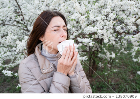 Woman blowing nose into tissue near white spring blossoms. Seasonal cold or allergy, hay fever symptom. Woman blowing nose into tissue near white spring blossoms. Seasonal cold or allergy, hay fever symptom. 139070909
