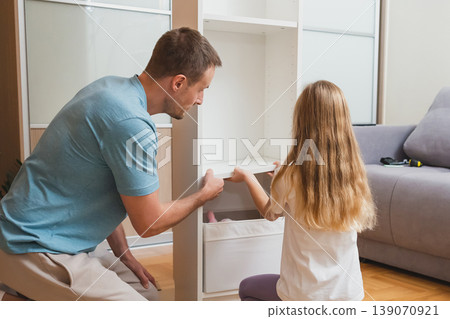 A father and daughter work together to place a white drawer into their newly assembled shelf in a bright living room with a gray sofa in the background A father and daughter work together to place a white drawer into their newly assembled shelf in a bright living room with a gray sofa in the background 139070921