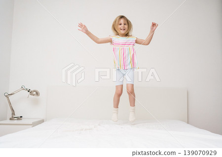 A smiling girl jumps on a bed with arms spread out in a bright minimalist room wearing a colorful rainbow striped shirt joyful energetic childhood play home family lifestyle concept A smiling girl jumps on a bed with arms spread out in a bright minimalist room wearing a colorful rainbow striped shirt joyful energetic childhood play home family lifestyle concept 139070929