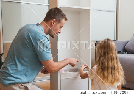 A father and daughter work together to place a white drawer into their newly assembled shelf in a bright living room with a gray sofa in the background 139070931