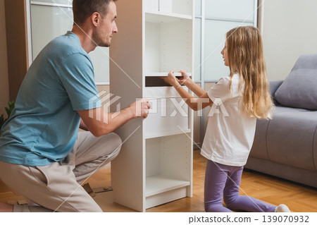 A father and daughter work together to place a white drawer into their newly assembled shelf in a bright living room with a gray sofa in the background 139070932