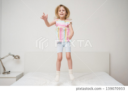 A smiling girl jumps on a bed with arms spread out in a bright minimalist room wearing a colorful rainbow striped shirt joyful energetic childhood play home family lifestyle concept 139070933