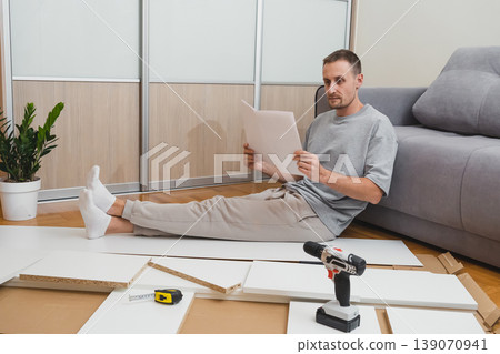 man sits on the floor reading furniture assembly instructions with tools and flat-pack parts scattered around him in a bright, modern living room man sits on the floor reading furniture assembly instructions with tools and flat-pack parts scattered around him in a bright, modern living room 139070941