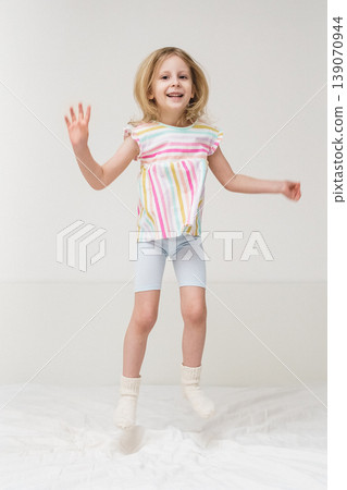 A smiling girl jumps on a bed with arms spread out in a bright minimalist room wearing a colorful rainbow striped shirt joyful energetic childhood play home family lifestyle concept 139070944