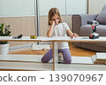 young girl sits among furniture parts on the floor, studying assembly instructions with tools and a screwdriver laid out before her in a bright living room 139070967
