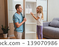 smiling father and daughter place white box on the newly assembled shelf in their living room, surrounded by tools and a gray sofa 139070986