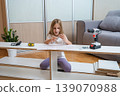 young girl sits among furniture parts on the floor, studying assembly instructions with tools and a screwdriver laid out before her in a bright living room 139070988