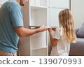 A father and daughter work together to place a white drawer into their newly assembled shelf in a bright living room with a gray sofa in the background 139070993