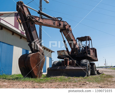 Weathered wheeled excavator stands idle near a striped building on a sunny day Weathered wheeled excavator stands idle near a striped building on a sunny day 139071035