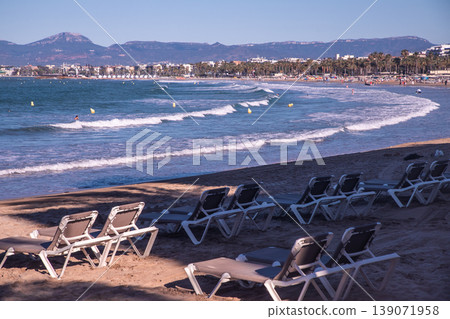 Beach scene with empty lounge chairs on sandy shore, gentle waves lapping at the coast and distant mountains under clear blue sky 139071958