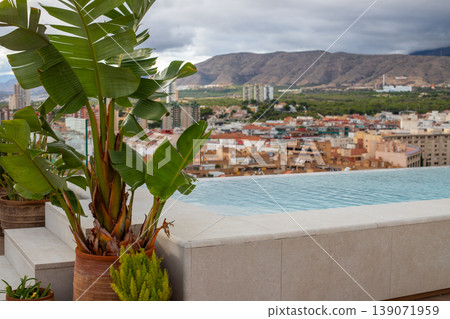 Rooftop infinity pool with tropical plants overlooking a cityscape and mountains under a cloudy sky in a Mediterranean region 139071959