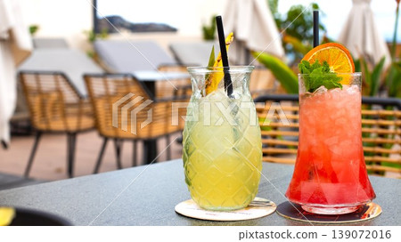Two colorful cocktails on a table in an outdoor cafe setting with lounge chairs and greenery in the background Two colorful cocktails on a table in an outdoor cafe setting with lounge chairs and greenery in the background 139072016