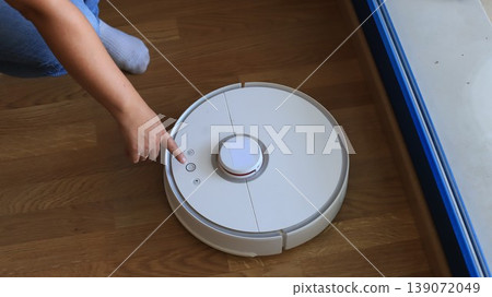 Person using a robotic vacuum cleaner on wooden floor, pointing at control buttons near a window with natural light streaming in Person using a robotic vacuum cleaner on wooden floor, pointing at control buttons near a window with natural light streaming in 139072049