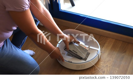 Woman removes dustbin from robotic vacuum cleaner while kneeling on wooden floor near a window with blue frame and sunlight streaming in Woman removes dustbin from robotic vacuum cleaner while kneeling on wooden floor near a window with blue frame and sunlight streaming in 139072050
