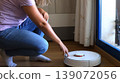Woman in casual attire interacts with a robotic vacuum cleaner on wooden floor near a window, showcasing modern home cleaning technology 139072056