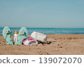 Beach scene with turquoise flip flops, sunscreen bottle, white sun hat, and rolled towel on sandy shore near calm blue ocean under clear sky 139072070