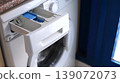Close-up view of a washing machine detergent drawer with blue compartment, set against a modern kitchen backdrop featuring a window and countertop 139072073