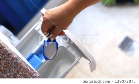 Hand holding a blue measuring scoop filled with detergent above a washing machine compartment in a bright laundry room setting Hand holding a blue measuring scoop filled with detergent above a washing machine compartment in a bright laundry room setting 139072075