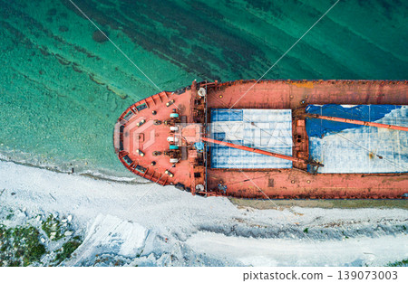 An aerial view shows a large, abandoned red cargo ship beached on a white pebble coastline. An aerial view shows a large, abandoned red cargo ship beached on a white pebble coastline. 139073003